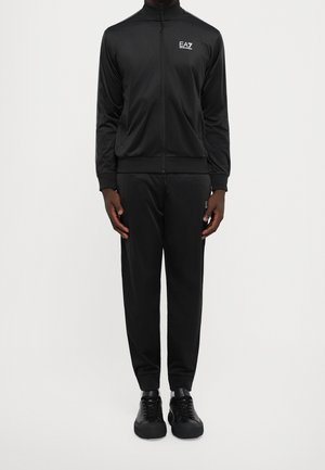 Mann steht und trägt eine schwarze EA7 Emporio Armani Zip-Up-Jacke, passende Hose und schwarze Sneaker vor weißem Hintergrund.