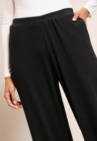 Melnas ribotās plaša kāju bikses ar teksturētu apdari, ar elastīgu jostu un sānu kabatām. Tuvplāns uz jostu un kāju.