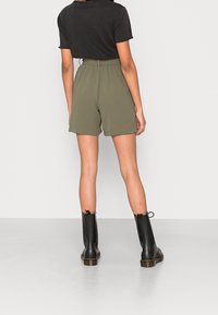 Pantaloni corti verdi oliva a vita alta con vestibilità ampia, abbinati a una maglietta nera a maniche corte e stivaletti neri, visti da dietro.