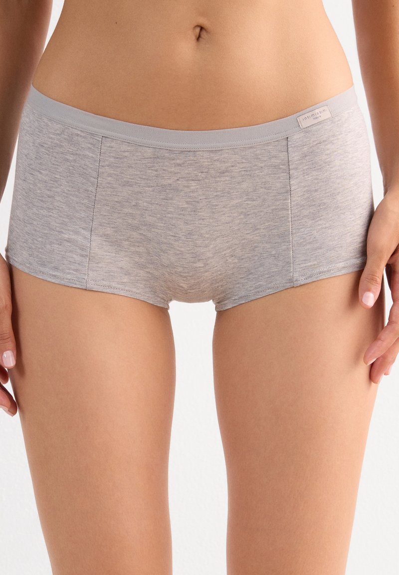 Mujer con ropa interior tipo boyshort de algodón gris claro con una pequeña etiqueta de marca en la cintura, posando sobre un fondo liso.