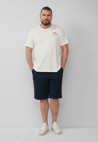 Hellbeige T-Shirt mit kurzen Ärmeln und Textakzenten, kombiniert mit marineblauen Shorts. Das Modell trägt weiße Sneakers und steht mit den Händen in den Taschen vor einem neutralen Hintergrund.