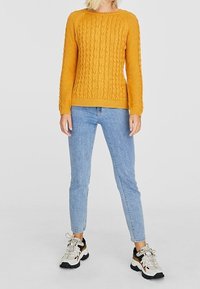 Pull tricoté jaune moutarde avec un motif en torsades texturé, associé à un jean skinny bleu clair et des baskets blanches épaisses.