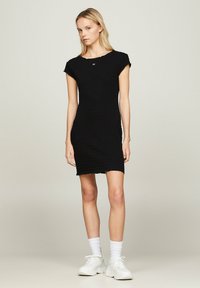 Vestido negro con textura acanalada, mangas cortas y un escote ligeramente deshilachado. Llevado con zapatillas blancas y calcetines. Diseño simple y ajustado.