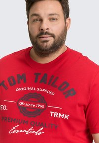 Rode katoenen T-shirt met zwarte tekst en een circulair logo, ronde hals en korte mouwen. Het ontwerp bevat "TOM TAILOR" en "sinds 1962."