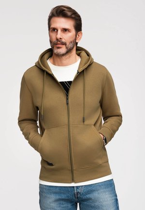 Hombre con barba que lleva una sudadera con capucha verde oliva con cremallera y vaqueros azules, de pie con las manos en los bolsillos contra un fondo claro y liso.