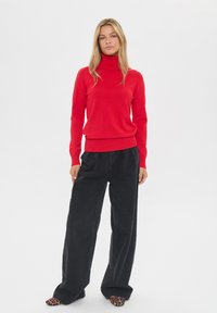 Crvena turtleneck džemper s rebrastim rubom, uparen s širokim crnim hlačama. Model nosi obuću s uzorkom leoparda, stoji ispred bijele pozadine.