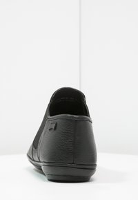 Zapato negro tipo slip-on con una textura de cuero suave, parte trasera redondeada y un diseño minimalista. Presenta un logo sutil en el talón.