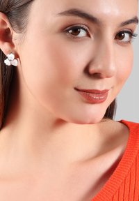 Silber Ohrringe in Form von drei sich überlappenden Herzen, mit einem strukturierten Design in einer hellen Farbe, getragen von einer Person in einem orangenen Pullover.