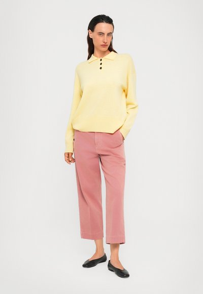 Woman wearing a pale yellow buttoned sweater, pink wide-leg cropped pants, and black flats, standing against a plain light background.