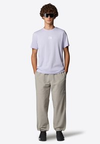 Jasnofioletowa koszulka z logo, beżowe lekkie spodnie i czarne buty. Model nosi duże czarne okulary przeciwsłoneczne. Prosty design i neutralne kolory.