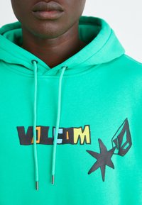 Grön hoodie med dragsko, som har en djärv svart och flerfärgad "VOLCOM"-logotyp samt en stjärngrafik på framsidan. Mjuk textur.