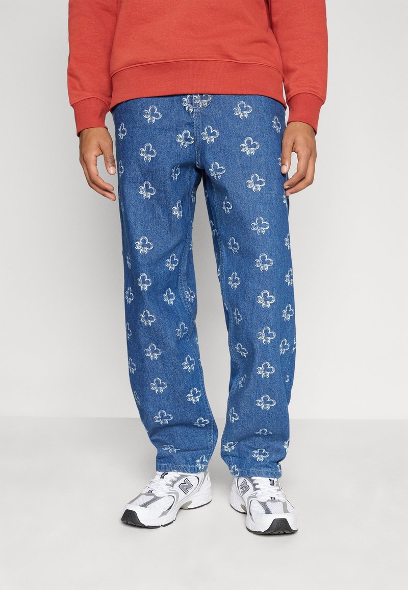 Homme portant un jean en denim bleu avec des motifs fleur-de-lis blancs, des baskets blanches et grises, et un sweat-shirt rouge.