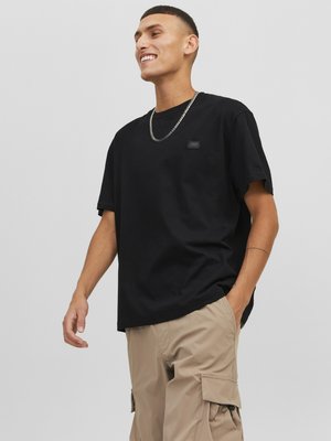 T-shirt en coton noir à manches courtes, coupe décontractée, avec un petit logo cousu sur la poitrine. Associé à un pantalon cargo beige.