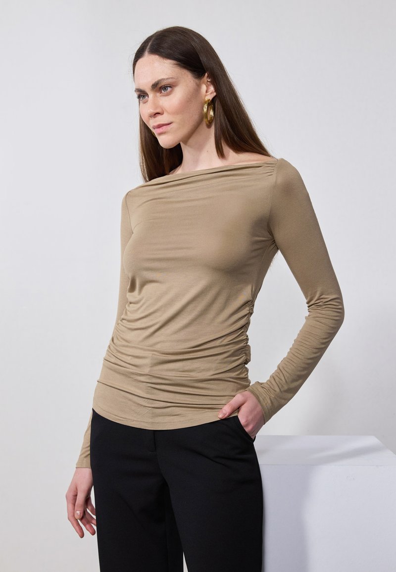 Beżowa bluzka z długim rękawem i dekoltem odkrywającym ramiona, z marszczonym wykończeniem w pasie, w połączeniu z czarnymi spodniami, na neutralnym tle.