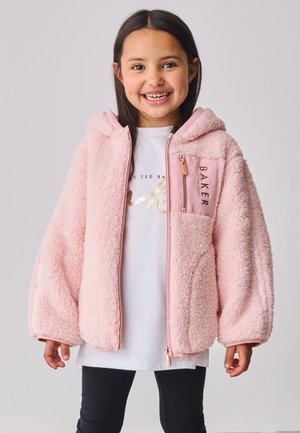 Fille souriante portant une grande veste à capuche douce et rose duveteuse par-dessus une chemise blanche et des leggings noirs, debout devant un fond clair uni.