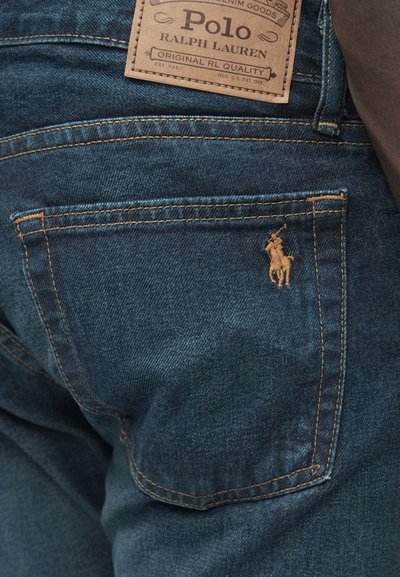 Tumši zili džinsa džinsi ar brūnas ādas etiķeti, uz kuras rakstīts "Polo Ralph Lauren". Aizmugurējā kabatā ir neliels izšūts zirga logotips.