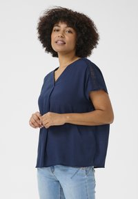 Blouse bleu marine à manches courtes avec un col en V. Tissu texturé et accents en dentelle sur les manches. Associée à un jean bleu clair.