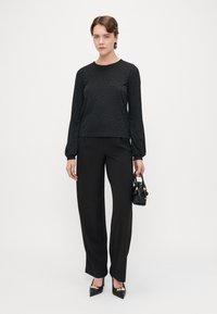 Top preto de manga longa com um padrão texturizado, combinado com calças pretas de cintura alta e sapatos de salto agudo pretos, segurando uma pequena mala.