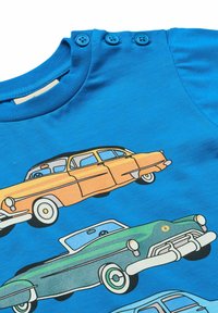 Plava pamučna majica s grafikom šarenih vintage automobila u narančastoj, zelenoj i svijetloplavoj boji. Ima tri plava gumba na izrezu.