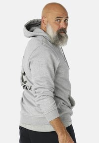 Sivi dukserica s kapuljačom s kenguruvim džepom, rebrastim manžetama i rubom. Na leđima se nalazi otisnuta oznaka. Tkanina izgleda mekano i teksturirano.