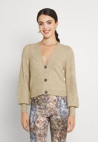 Beige geribbelde cardigan met een diepe V-hals, vier donkere knopen en pofmouwen, gecombineerd met gestreepte leggings.