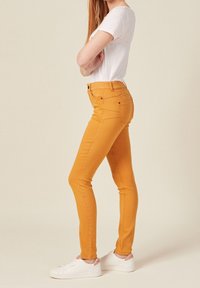 Slim-fit yellow jeans made from denim, featuring five pockets, belt loops, and a mid-rise waist. Paired with white sneakers.