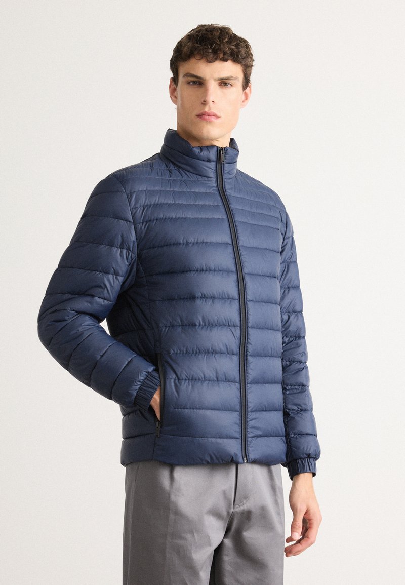 Young man with curly hair wearing a zipped navy blue quilted jacket and gray pants, one hand in jacket pocket, standing against a white background.