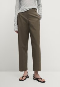 Personne portant un pantalon droit olive, un haut à manches longues gris clair et des sandales plates noires à lanières se tenant devant un fond uni.