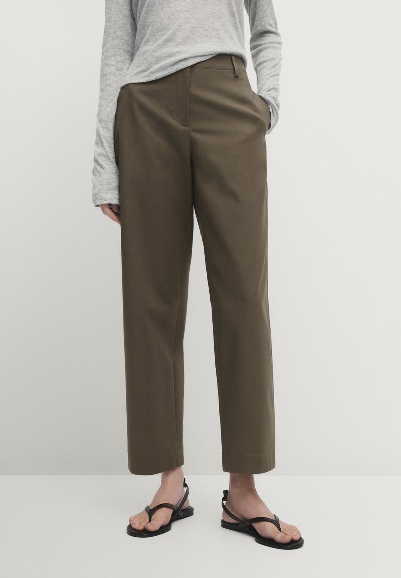 Personne portant un pantalon droit olive, un haut à manches longues gris clair et des sandales plates noires à lanières se tenant devant un fond uni.