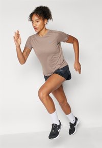 Lys brun idretts t-skjorte med korte ermer og en subtil logo, kombinert med svarte shorts og svarte joggesko, med hvite sokker.