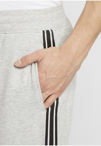 Handen vilar i fickan på ljusgråa sweatpants med svart och vita vertikala ränder längs sidsömmen.