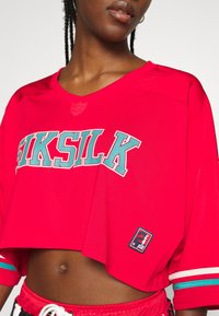 Rotes, kurzes Jersey mit "SIKSILK" in türkisfarbener Schrift, strukturiertem Stoff, kurzen Ärmeln und einem Logo-Patch am Saum. Weiße und türkisfarbene Akzente.