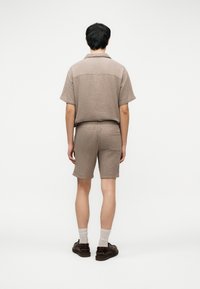 Άτομο που στέκεται με την πλάτη, φορώντας μπεζ υφασμάτινο κοντομάνικο πουκάμισο και ταιριαστά σορτς, λευκές κάλτσες και σκούρα καφέ παπούτσια.