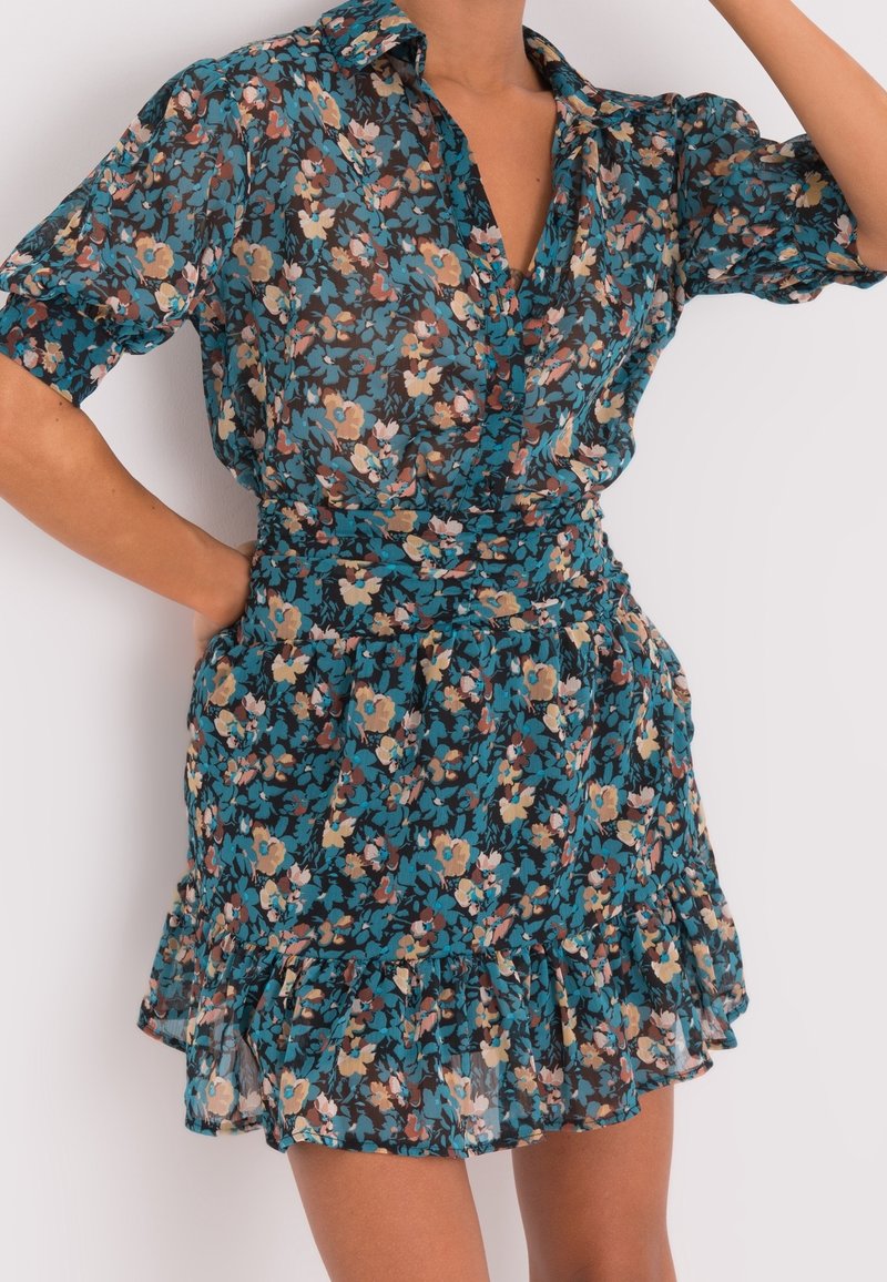 Kukkamekko läpinäkyvästä kankaasta, jossa on tumma sininen tausta ja monivärisiä kukkia, lyhyet hihat, kapea vyötärö ja röyhelöhelma.