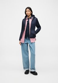 Γυναίκα που φοράει μια μπλε σκούρα καπιτονέ μπουφάν πάνω από ροζ πουκάμισο και μαύρο τοπ, ανοιχτό μπλε τζιν με πλατύ μπατζάκι και μαύρα παπούτσια με πλατφόρμα, στέκεται με το χέρι στην τσέπη.