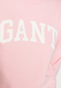 Sweat-shirt rose clair avec un col rond et des lettres blanches en relief "GANT" sur la poitrine. Tissu doux et texture lisse.