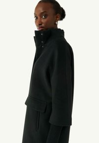 Cappotto di lana nero con colletto alto, dettagli con bottoni e tasche laterali. Presenta polsini a coste e una silhouette strutturata.
