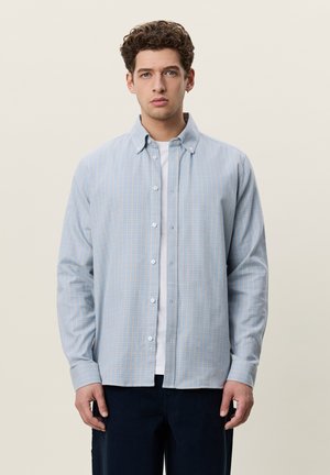 Jeune homme aux cheveux bouclés portant une chemise à carreaux bleu clair boutonnée par-dessus un t-shirt blanc, debout devant un fond uni.