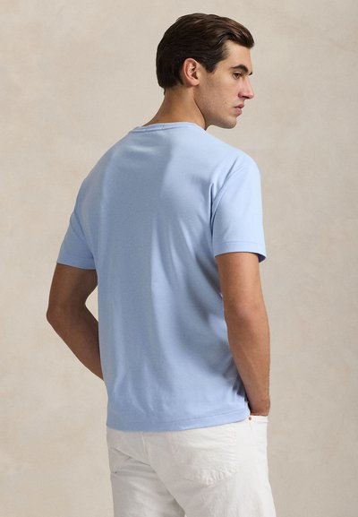 Šviesiai mėlyna trumpomis rankovėmis marškinėlių su švelnios medvilnės audiniu, su apvalia iškirpte ir laisvu kirpimu, žiūrint iš nugaros prieš neutralų foną.