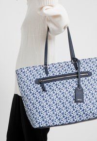 Modrý a bílý taška přes rameno s koženými doplňky, přihrádkou na zip a dvěma uchy, zobrazující geometrický vzor na plátěném materiálu.