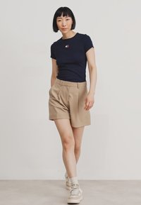 Mörkblå t-shirt med kort ärm och en liten broderad design, som kombineras med skräddarsydda beige shorts. Skorna har en ljusbeige och vit design.