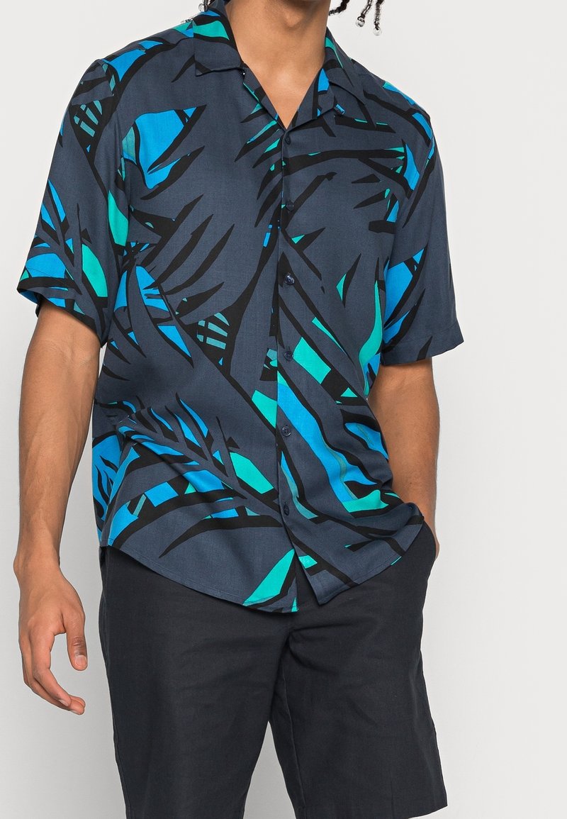 Lyhythihainen tummansininen paita, jossa rohkea musta ja turkoosi lehtikuvio, kevyt kangas ja etupuolelta napillinen malli.