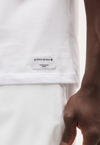 Λευκή ετικέτα μπλουζάκι Björn Borg κοντά στο τελείωμα, φορεμένη από άτομο με σκουρόχρωμο δέρμα, δίπλα σε λευκό παντελόνι και ορατό δεξί χέρι.