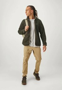 Olivno zelena flis jakna, na zadrgo, v kombinaciji s svetlo sivo majico in bež vojaškimi hlačami. Nositi lahkoten nahrbtnik in temne čevlje.