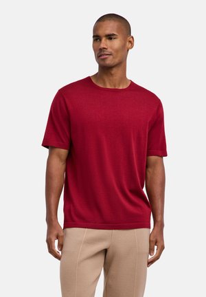 Uomo con indosso una maglietta rossa a maniche corte e pantaloni beige, in piedi davanti a uno sfondo chiaro e uniforme, che guarda leggermente verso destra.