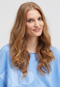 Light blue oversized sweatshirt with a crew neck and textured fabric. Model has wavy, shoulder-length hair and a subtle smile.