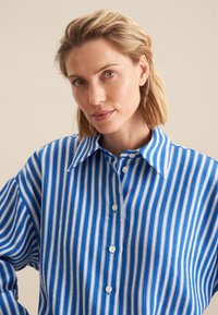 Blue and white striped button-up shirt with a classic collar and relaxed fit. Material appears lightweight with a smooth texture.