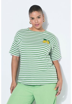 Henkilö, jolla on vihreä-valkoraidallinen paita, jossa on sitruunapala, ja vaaleanvihreät housut, seisoo valkoista taustaa vasten.