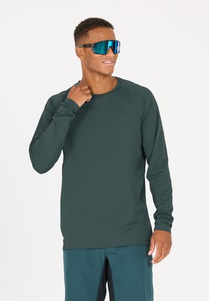 Langärmliges Hemd aus dunkelblauem Stoff mit einem strukturierten Muster. Modell trägt blaue verspiegelte Sonnenbrille und nimmt eine lässige Pose ein.