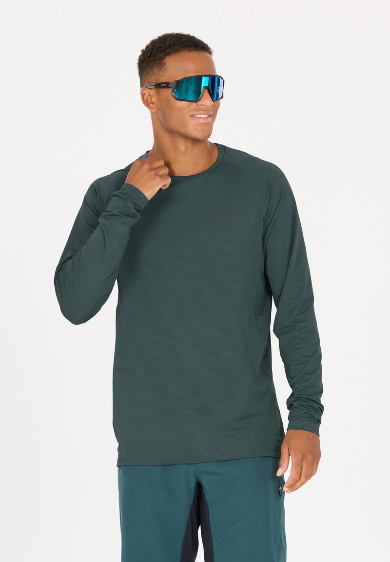 Långärmad skjorta i mörk teal-tyg med ett texturerat mönster. Modell som bär blå spegelglasögon, ger en avslappnad pose.